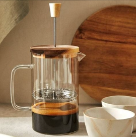 Clear glass French press with wooden lid and handle, filled with coffee, on a neutral background.